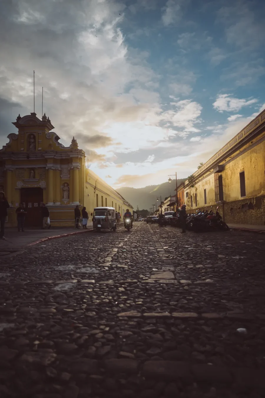 Sunset on a cobblestone street in Antigua Guatemala with the yellow façade of the Capilla de Belén and a tuk tuk approaching