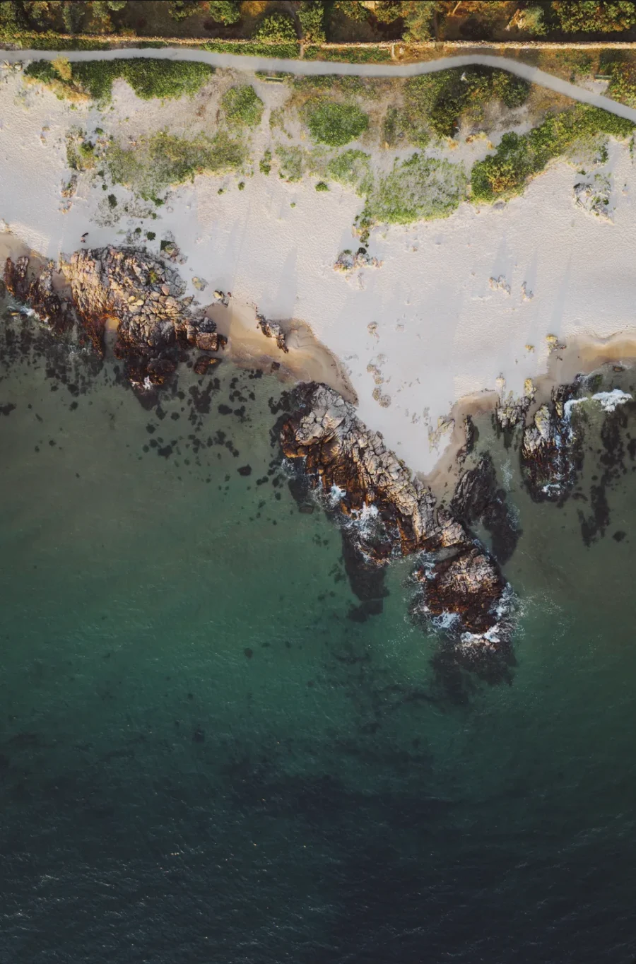 Looking straight down at Sandkås beach on Bornholm at sunrise, where pale sand, a spine of rock and clear Baltic water sort into bands