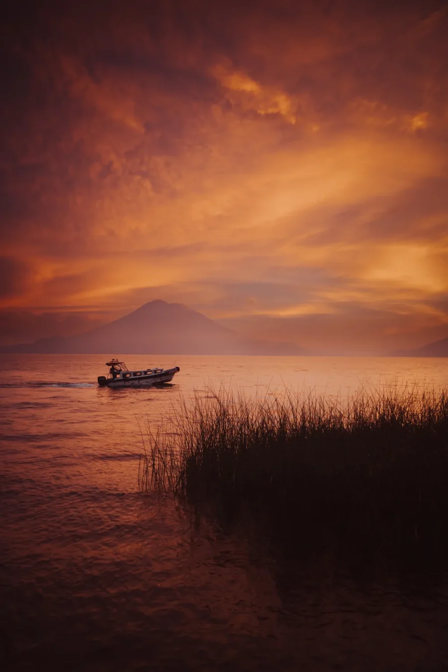 A boat crossing Lake Atitlán at sunset under a burning orange sky, with a volcano silhouetted across the water and reeds in the foreground