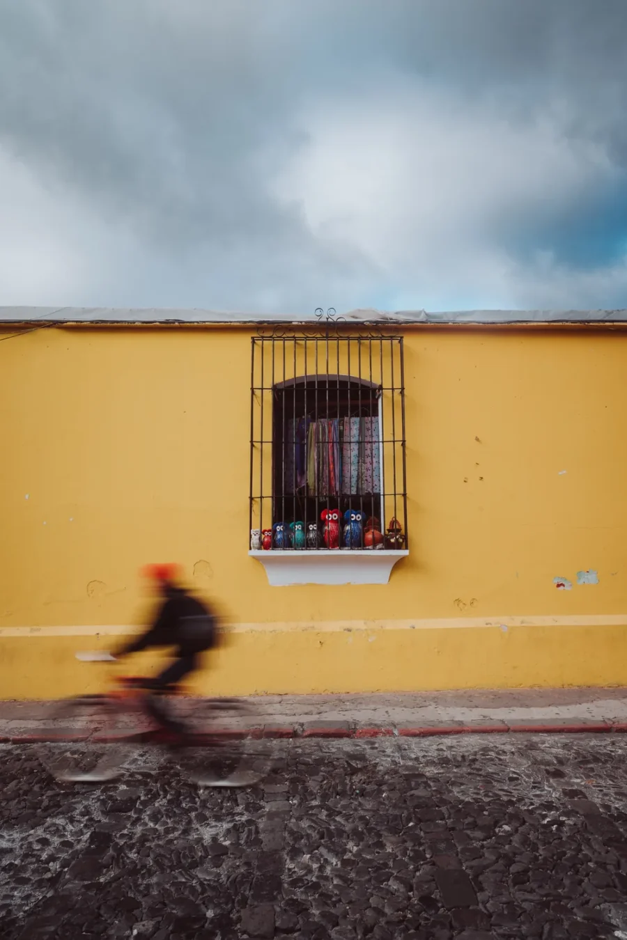 A row of small crochet owls behind window bars on a yellow market wall in Antigua Guatemala with a blurred cyclist passing