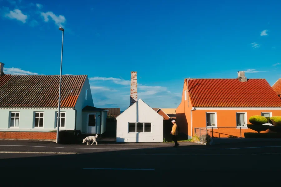 A woman walking a white dog past mint green, white and orange houses on an empty street in Tejn, Bornholm, Denmark, in low evening sun