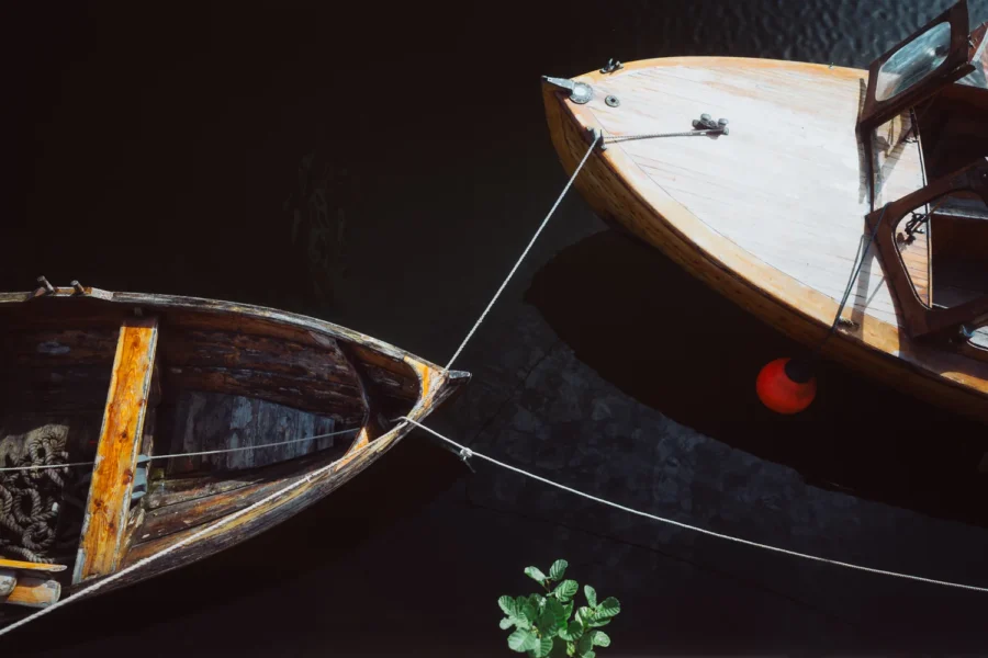 Two old wooden boats tied bow to bow on black water in Bjørvika, Oslo, with a single red buoy and a green sprig the only colour in the frame