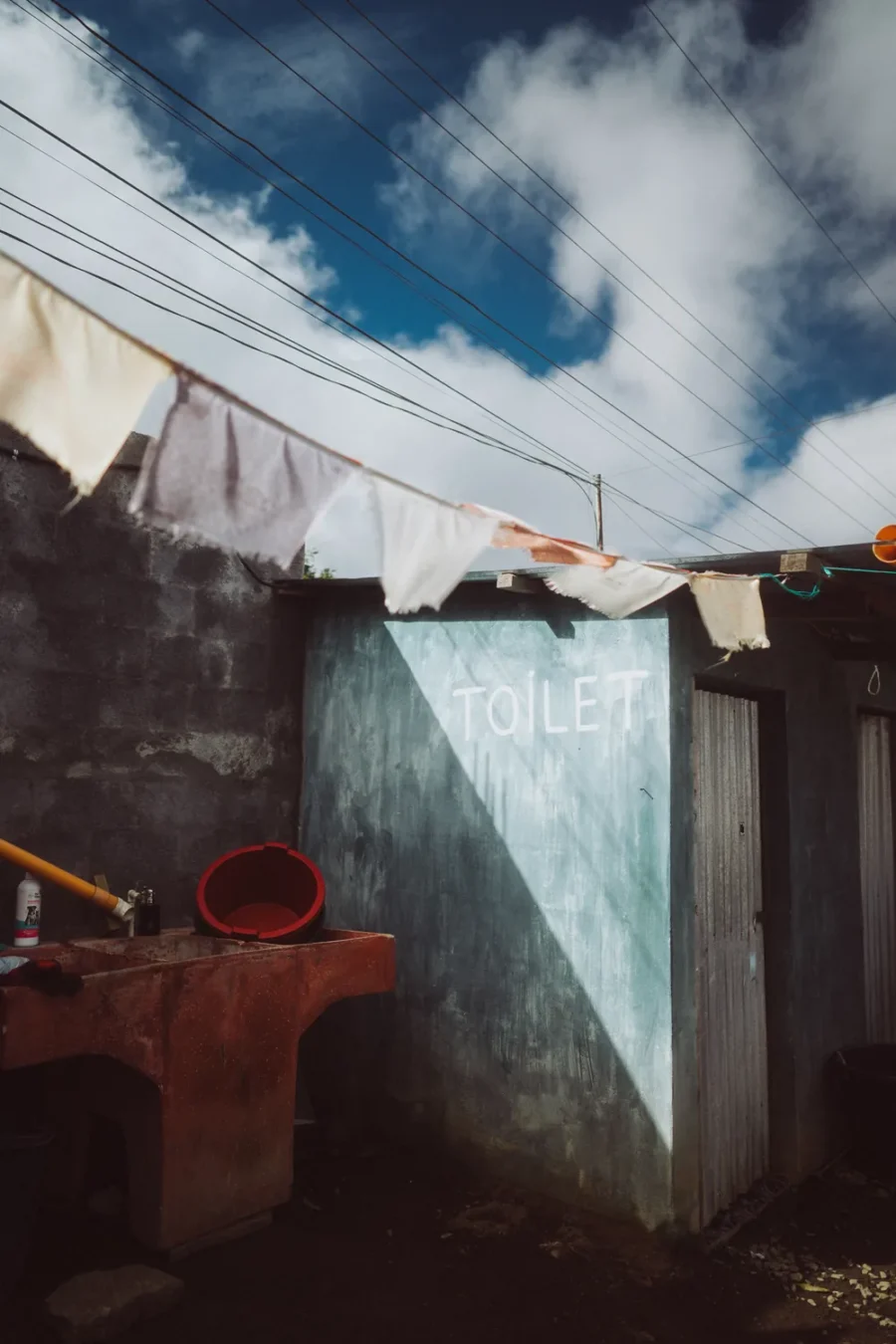 A concrete washblock with the word TOILET painted on it, hard midday shadow and a clothesline of rags under a deep blue sky, Guatemala