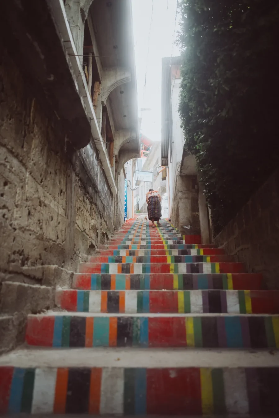 A narrow flight of rainbow painted steps climbing between two buildings in San Pedro La Laguna, Guatemala, with a woman and a dog ascending