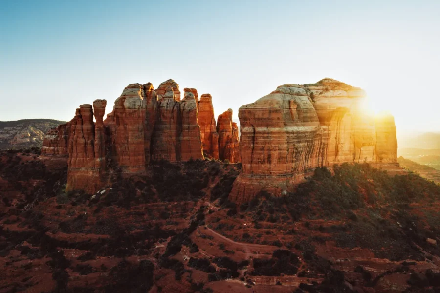 Aerial view of Cathedral Rock in Sedona, Arizona at sunset, the sun flaring off the right spire above red rock and green desert brush.
