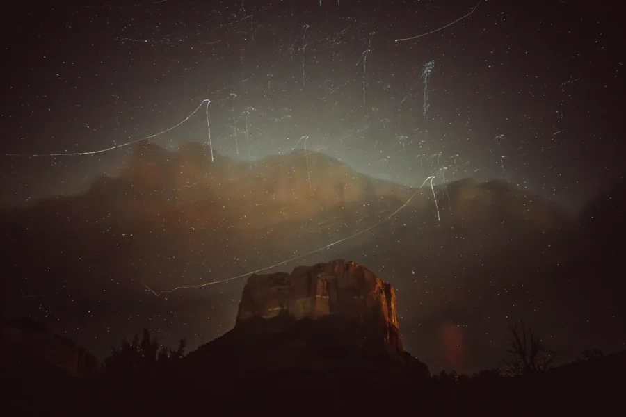 Night photograph of Courthouse Butte in Sedona, Arizona, with a ghostly image of a distant mountain range and streaked stars layered in the sky above it.