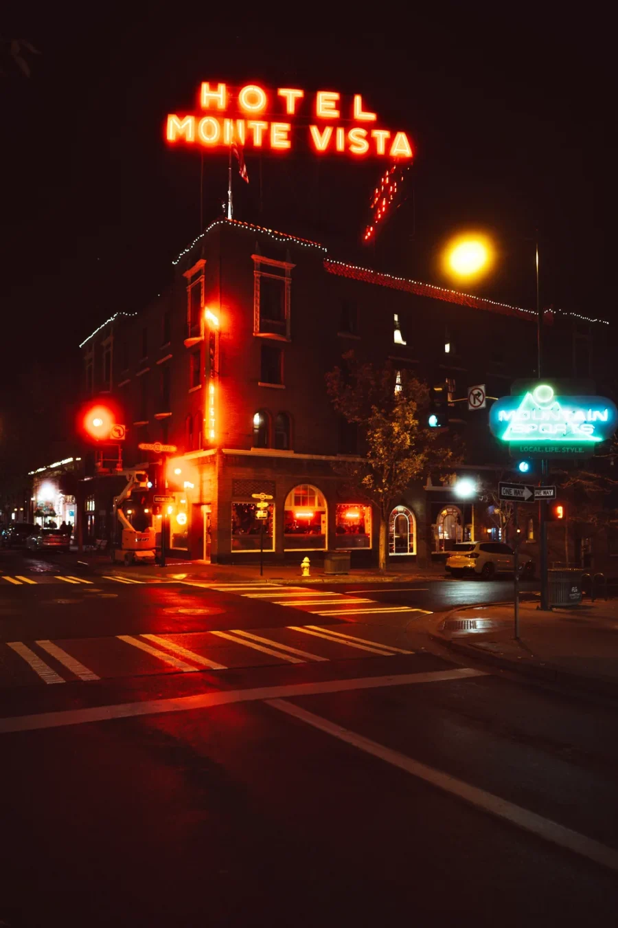 Hotel Monte Vista's red neon rooftop sign glowing over a rain-wet street corner in downtown Flagstaff, Arizona at night, with a green Mountain Sports sign at right.