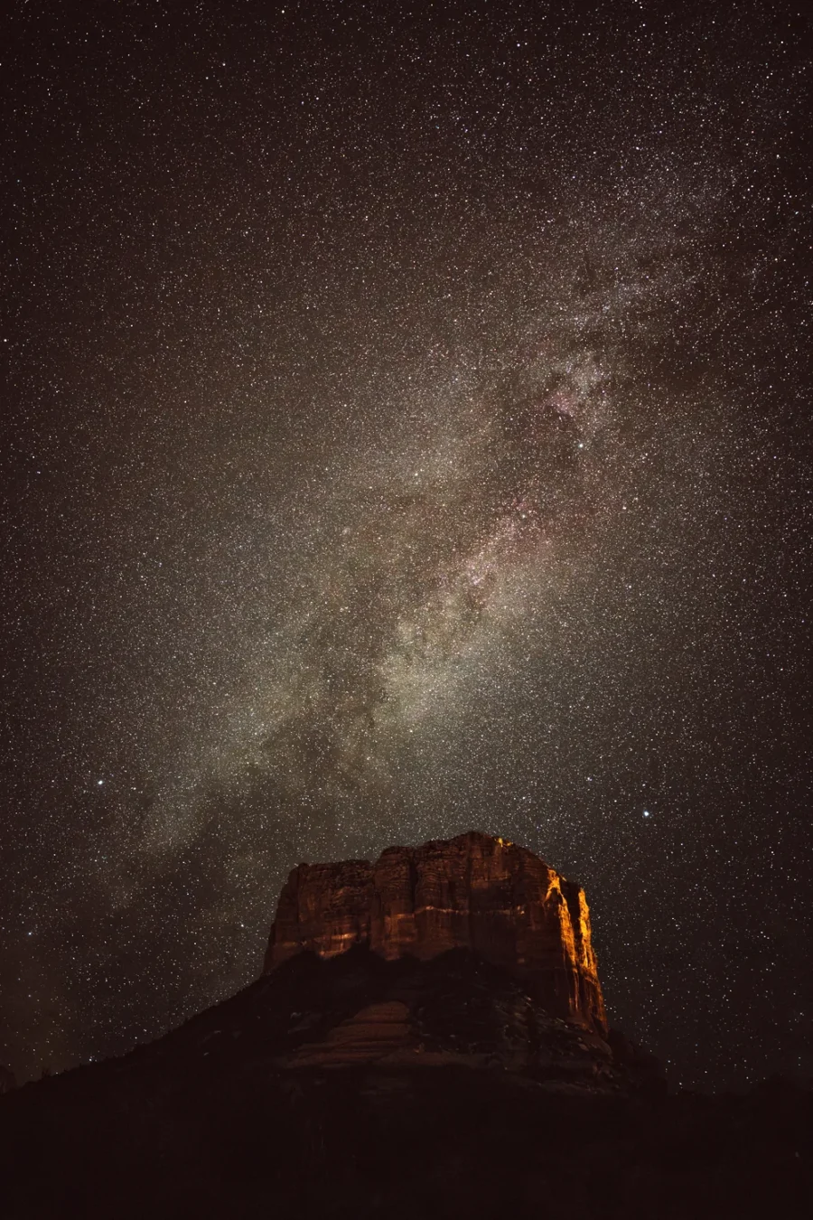 Milky Way rising through a star-filled sky above Courthouse Butte in Sedona, Arizona, the butte's face lit warm orange by distant town light.