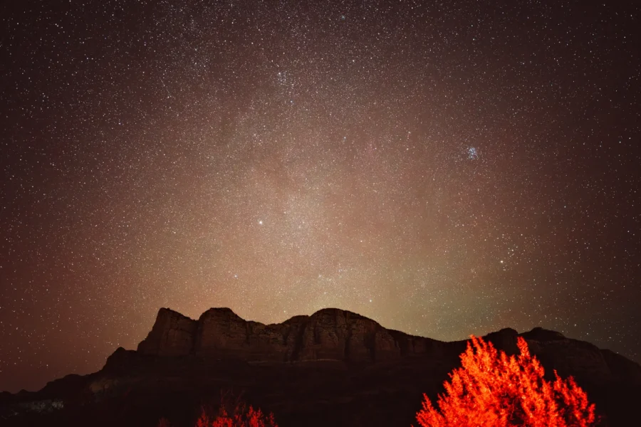Star-filled night sky over a red rock ridge near Courthouse Butte in Sedona, Arizona, with the Pleiades star cluster at upper right and a tree glowing red in the foreground.
